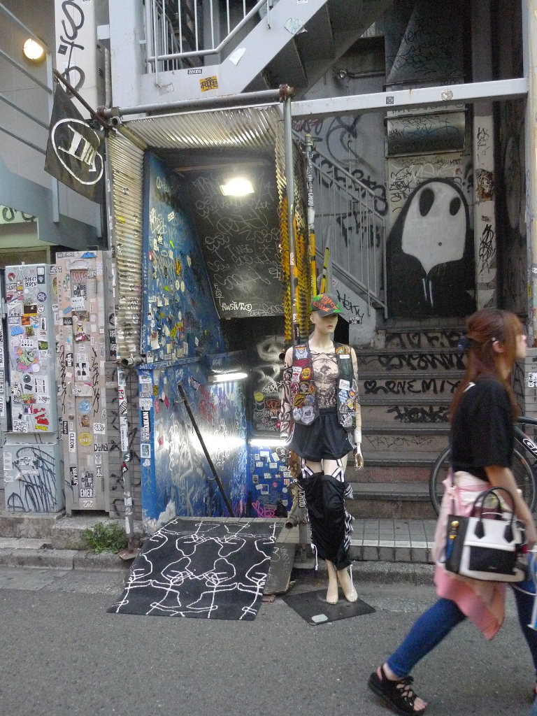 Un mannequin, devant un mur de graffitis, semble regarder une passante.